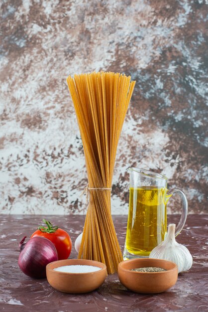 Espaguetis crudos, aceite de oliva y verduras frescas en la superficie de mármol.