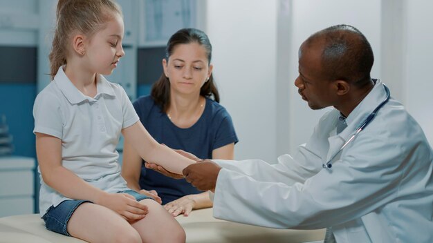 Especialista afroamericano que examina la lesión de un niño pequeño en el gabinete. Médico general consultando a una niña pequeña con dolor en el brazo fracturado, brindando asistencia y apoyo médico.