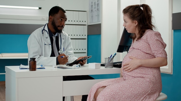 Especialista en maternidad tomando notas en tabletas digitales en el chequeo con mujeres embarazadas para dar medicamentos recetados. Médico varón hablando con paciente con barriga de embarazo en la cita.