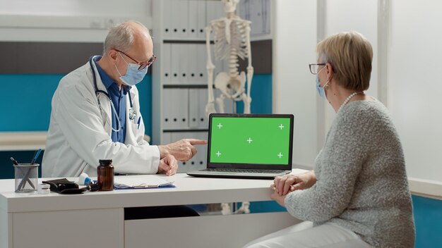 Especialista médico que muestra pantalla verde en la computadora portátil a la mujer en el chequeo en el gabinete. Mirando el fondo del espacio de copia en blanco con una plantilla de maqueta aislada y una clave de croma. Disparo de trípode.