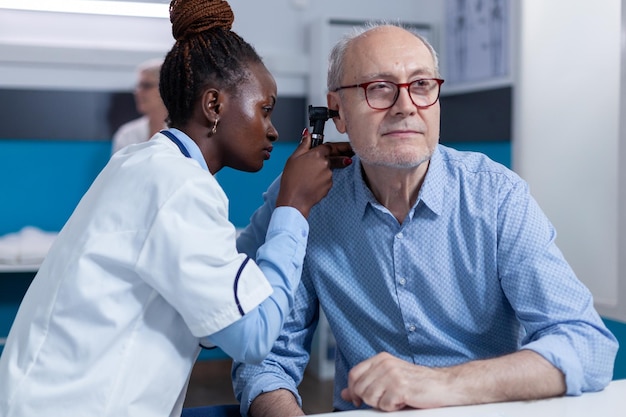 Especialista en otología clínica que consulta a pacientes mayores que usan otoscopio para controlar la infección del oído. El otólogo del hospital examina la condición del oído interno del jubilado enfermo mientras está en el gabinete del médico.