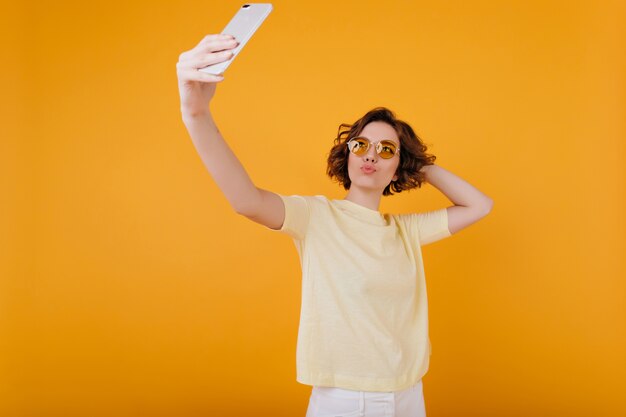 Espectacular chica caucásica con cabello castaño haciendo selfie en habitación amarilla