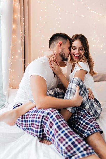 Espectacular chica en pantalones a cuadros posando juguetonamente en la cama con su marido. Chico de buen humor abrazando a su esposa por la mañana.