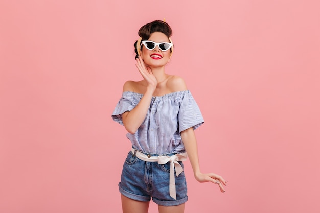 Espectacular chica pinup en gafas de sol sonriendo a la cámara. Foto de estudio de mujer bonita estilizada aislada sobre fondo rosa.