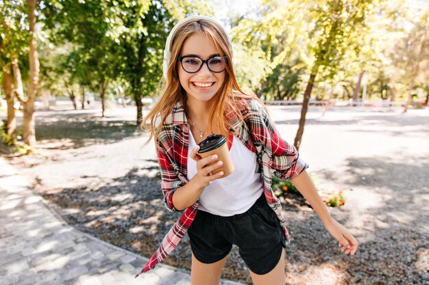 Espectacular chica en shorts negros bailando en el parque y bebiendo café Disparo al aire libre de mujer feliz riendo con cabello rubio