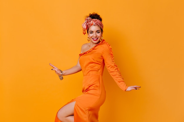 Espectacular dama con diadema inusual y vestido de satén con abertura está sonriendo en el espacio naranja.