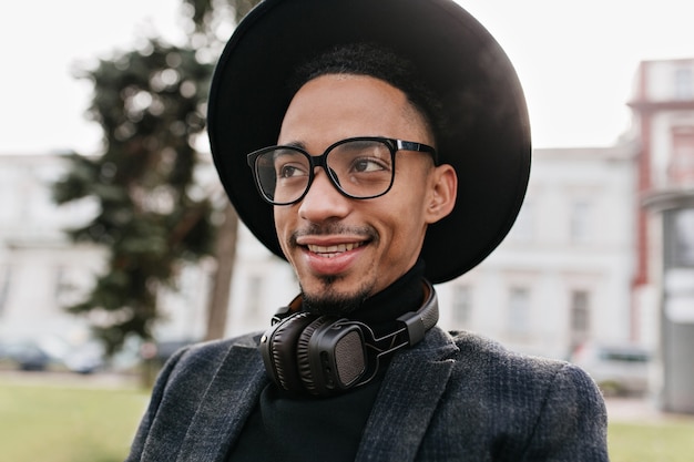 Espectacular hombre africano con sincera sonrisa posando. Foto al aire libre del lindo modelo masculino negro con gafas y auriculares.