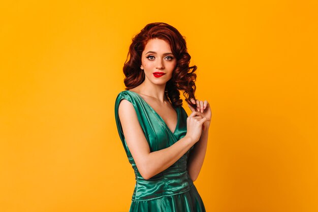 Espectacular joven tocando el pelo de jengibre rizado en el espacio amarillo. Foto de estudio de increíble modelo femenino en vestido verde.