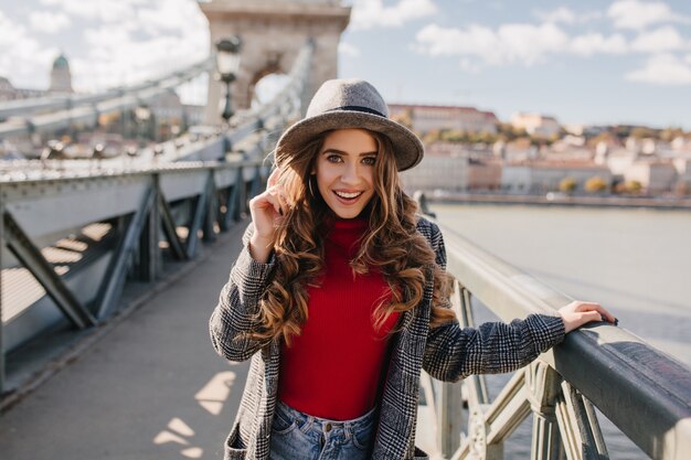 Espectacular modelo femenino blanco con sombrero posando sobre fondo de río y sonriendo