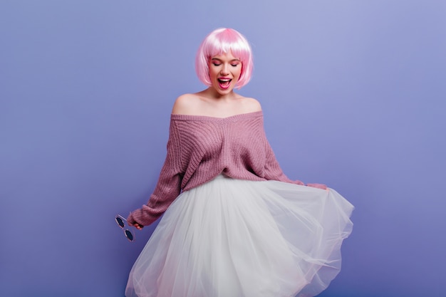 Espectacular modelo de mujer caucásica en corto periwig bailando en la pared púrpura. Foto interior de una encantadora dama blanca con cabello rosado posando en una falda larga y exuberante.