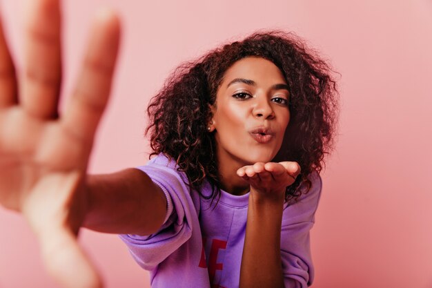 Espectacular mujer africana expresando amor mientras hace selfie. Chica rizada optimista tomando una foto de sí misma.