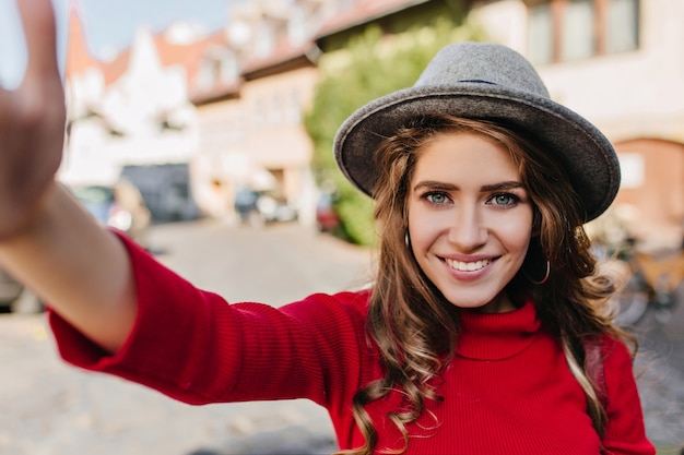 Espectacular mujer blanca de ojos azules con elegante sombrero haciendo selfie con sonrisa dichosa