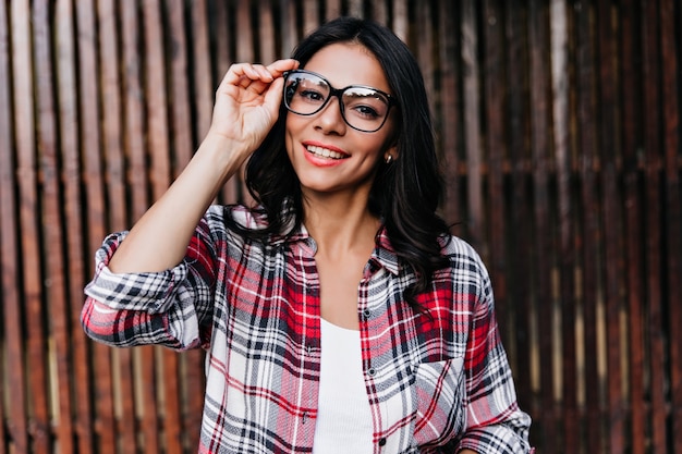 Espectacular mujer bronceada tocando juguetonamente sus gafas en la pared de madera. Encantadora dama con peinado ondulado sonriendo.