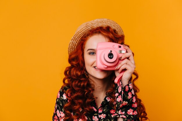 Espectacular mujer con cabello rojo ondulado y ojos azules cubre su rostro, tomando fotos con mini cámara.
