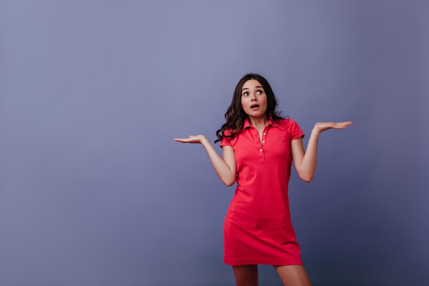 Espectacular mujer joven en traje rojo posando con las manos en alto. Chica de pelo castaño sorprendida de pie en la pared púrpura con la boca abierta.