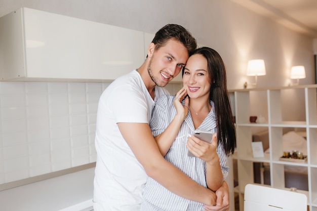 Espectacular mujer morena con celular en mano posando en los brazos del marido con una sonrisa