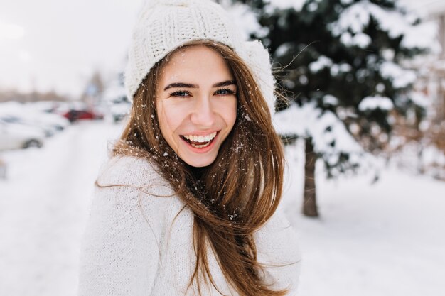 Espectacular mujer de pelo largo riendo mientras posa sobre la nieve. Foto de primer plano al aire libre de modelo de mujer caucásica con sonrisa romántica escalofriante en el parque en día de invierno.