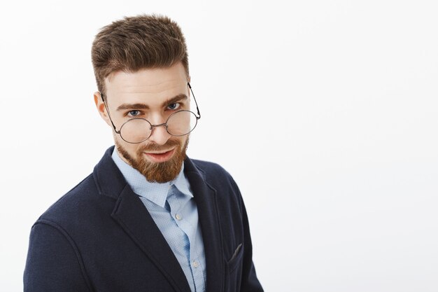 Espera de tiro de hombre europeo inteligente y exitoso encantador con barba y ojos azules mirando por debajo de las gafas con expresión sexy de pie en traje elegante posando contra la pared blanca