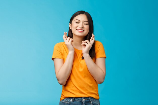 Esperanza emocionada joven asiática haciendo deseo cruzar los dedos buena suerte cerrar los ojos sonriendo creer soñar ...