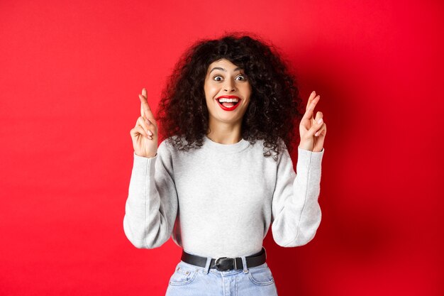 Esperanza emocionada mujer pidiendo un deseo, cruzar los dedos para la buena suerte y sonriendo asombrada a la cámara, rezando para que el sueño se haga realidad, esperando resultados, fondo rojo.