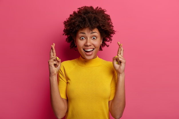 Foto gratuita esperanza emocionada mujer de piel oscura cree que el trato va bien, pide un deseo, cruza los dedos para tener buena suerte, suplica y ora, sonríe ampliamente, muestra dientes blancos, usa camiseta amarilla, pone todos los esfuerzos en orar