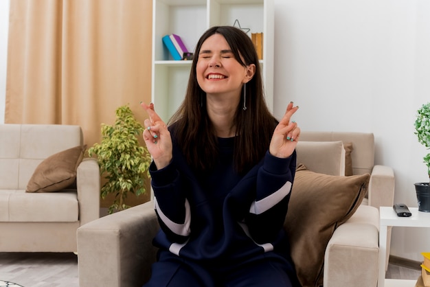 Esperanza joven bonita mujer caucásica sentada en un sillón en la sala de estar diseñada cruzando los dedos y deseando buena suerte con los ojos cerrados