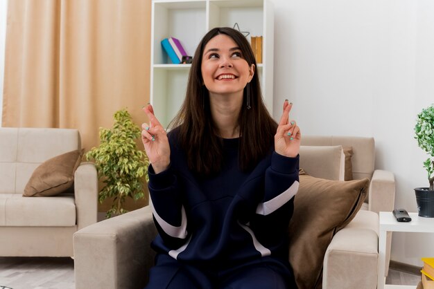 Esperanza joven bonita mujer caucásica sentada en un sillón en la sala de estar diseñada mirando al lado cruzando los dedos y deseando buena suerte