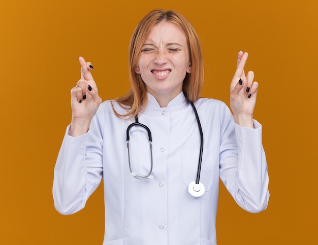 Esperanza joven médico de jengibre vistiendo bata médica y estetoscopio haciendo gesto de buena suerte con los ojos cerrados aislados en la pared naranja