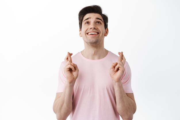 Foto gratuita esperanza y suerte un joven sonriente cruza los dedos y mira hacia arriba rezando pidiéndole a dios que haga realidad el deseo anticipando noticias positivas de pie sobre fondo blanco