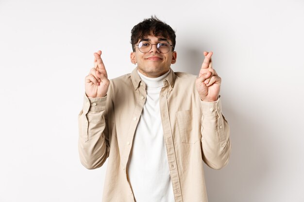 Esperar. Feliz joven pidiendo deseos y sonriendo, mirando esperanzado a la cámara mientras espera que el sueño se haga realidad, cruce los dedos para la buena suerte, de pie sobre fondo blanco.