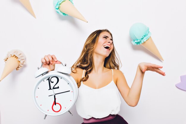 Espléndida mujer joven en camiseta blanca posando con los ojos cerrados sosteniendo el reloj en la pared decorada. Retrato de niña morena emocionada riendo de pie delante de la pared con dulces de juguete.