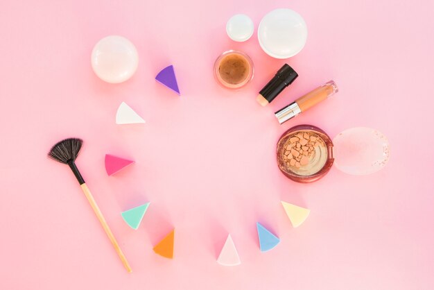 Esponjas cosméticas de diferentes colores con productos de maquillaje sobre fondo rosa.