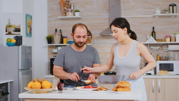 Esposa cocina huevos para su esposo durante el desayuno mientras él unta mantequilla en pan tostado. Usar pijamas por la mañana, preparar la comida juntos, la joven pareja feliz se ama y se casa