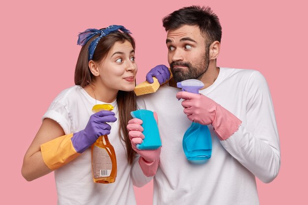 La esposa y el esposo europeos divertidos sostienen un trapeador y una botella de spray, usan guantes protectores de goma