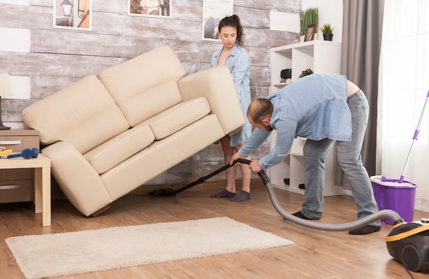 Foto gratuita esposa levanta el sofá mientras su esposo limpia el polvo debajo de él con una aspiradora