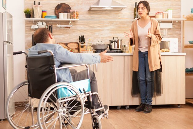 Esposa mirando enojada con un hombre discapacitado en silla de ruedas durante una discusión sobre su relación en la cocina. Hombre discapacitado paralítico discapacitado con discapacidad para caminar que se integra después de un accidente.