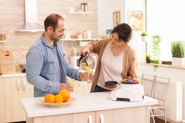 Esposa vertiendo sabroso batido mientras el marido sostiene el vaso. Estilo de vida saludable, despreocupado y alegre, comiendo dieta y preparando el desayuno en una acogedora mañana soleada