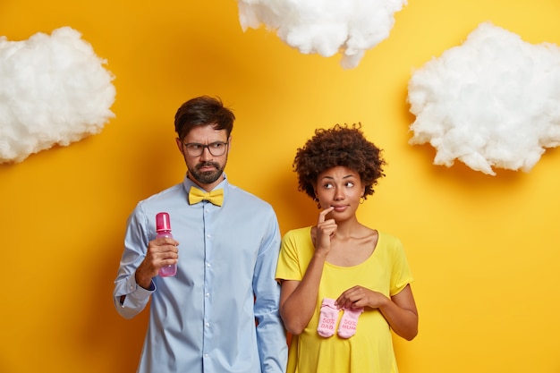 Foto gratuita el esposo y la esposa esperan al bebé, posan con biberón y botines de recién nacido, piensan en el nombre del futuro niño, prepárense para convertirse en padres, aislados en nubes amarillas y blancas sobre su cabeza