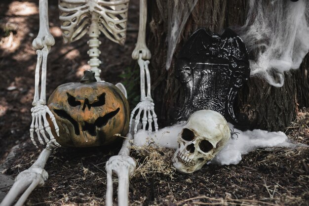 Esqueleto con calabaza sentada cerca de cráneo y lápida apoyándose en el árbol