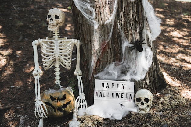 Esqueleto con calabaza sentada cerca de la tableta de Halloween apoyándose en el árbol