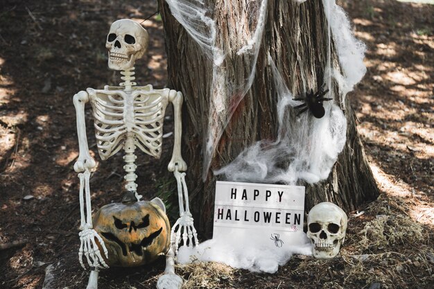 Esqueleto con calabaza sentada cerca de la tableta de Halloween apoyándose en el árbol