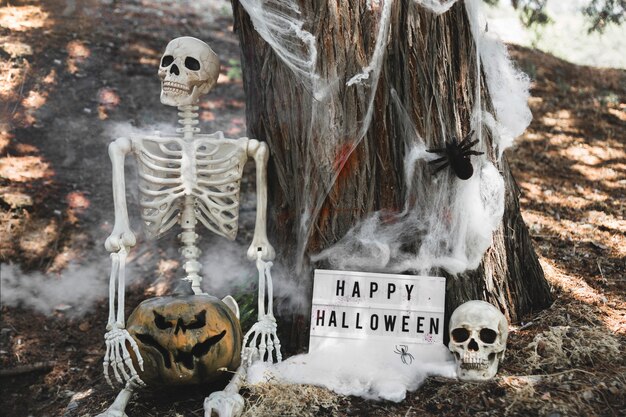 Esqueleto con calabaza sentado en la niebla cerca de la tableta apoyándose en el árbol