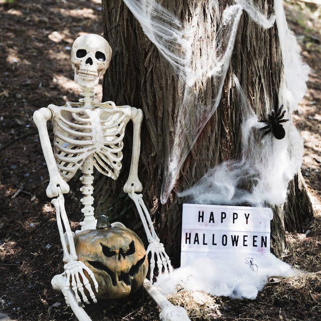 Esqueleto sentado cerca de árbol con calabaza y inscripción de Feliz Halloween