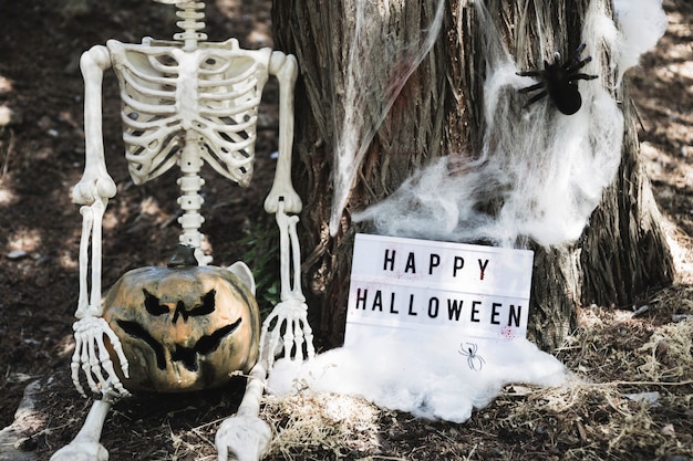 Esqueleto sentado cerca de la tableta de Halloween apoyándose en el árbol