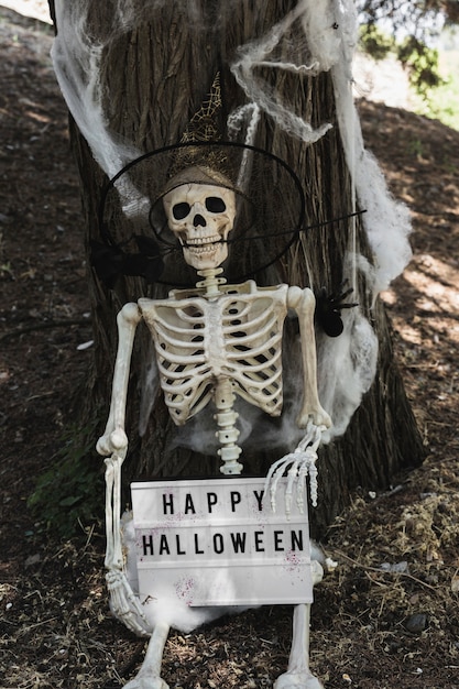 Esqueleto en sombrero de mago sosteniendo la tableta de Halloween cerca de árbol