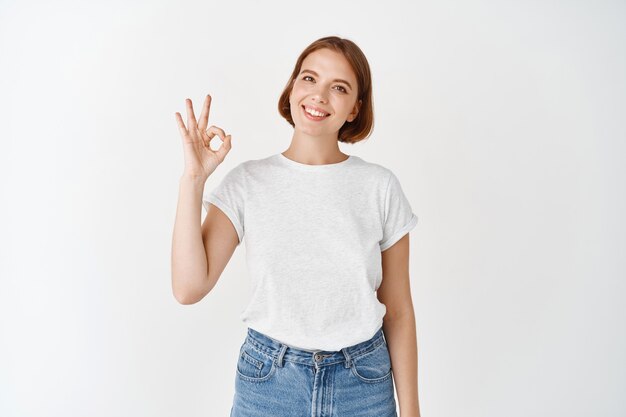 Está bien. Mujer joven sonriente que muestra un signo bien, aprobar o recomendar algo bueno, alabando la excelente calidad, de pie en la pared blanca