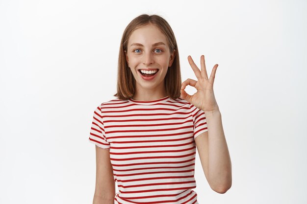 Está bien, sí. Una chica rubia sonriente y alegre confirma que todo está bajo control, muestra un gesto bien, bien cero y asiente con aprobación, dice sí, te alaba o te felicita, fondo blanco.