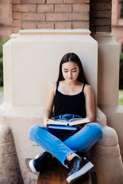 Foto gratuita estacione el libro de lectura de la mujer en el banco que sonríe feliz en la cámara. mujer multicultural bastante joven que disfruta de la primavera en parque.