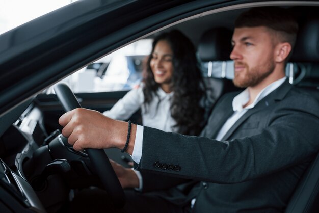 El estado de ánimo alegre. Encantadora pareja exitosa probando coche nuevo en el salón del automóvil