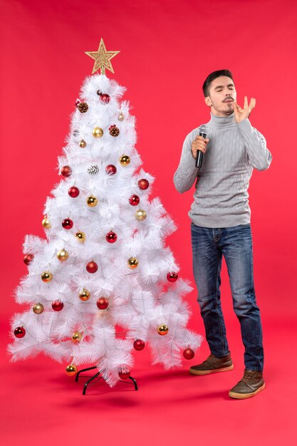 Estado de ánimo de año nuevo con chico positivo vestido con jeans de pie cerca del árbol de Navidad decorado